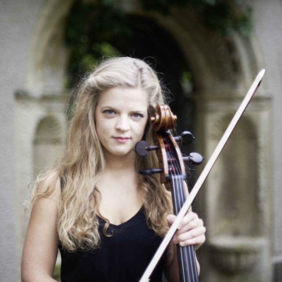 Die Cellistin Alma Stolte steht mit Ihrem Cello in einem schwarzen Kleid vor einem Torbogen