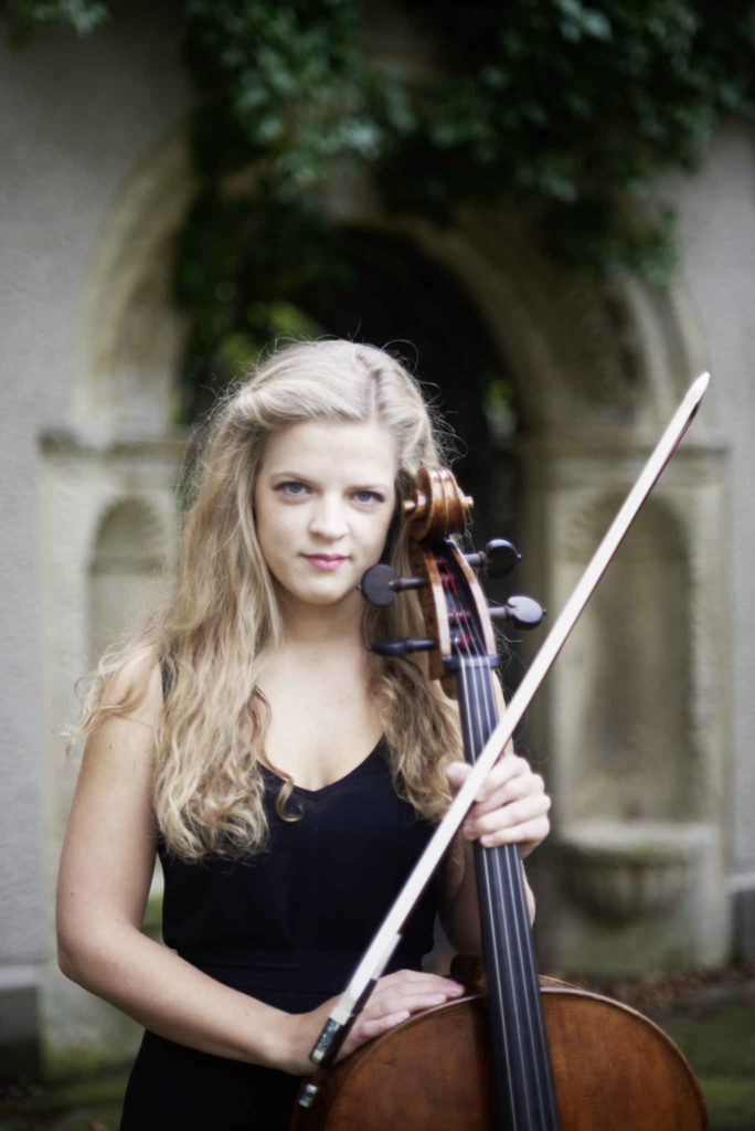 Die Cellistin Alma Stolte steht mit Ihrem Cello in einem schwarzen Kleid vor einem Torbogen
