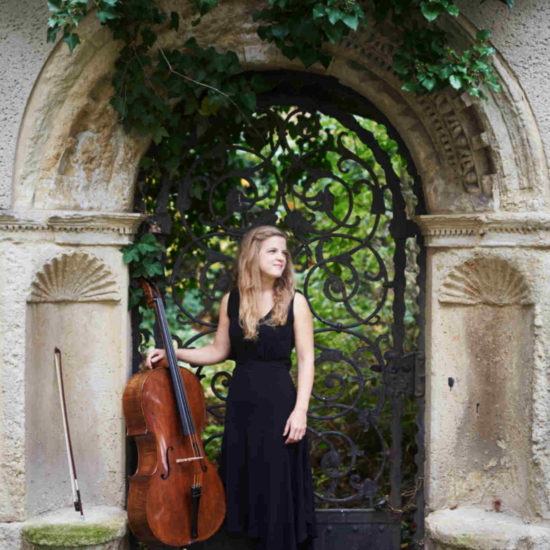 Die Cellistin Alma Stolte steht neben ihrem Cello in einem schwarzen Kleid in einem Torbogen