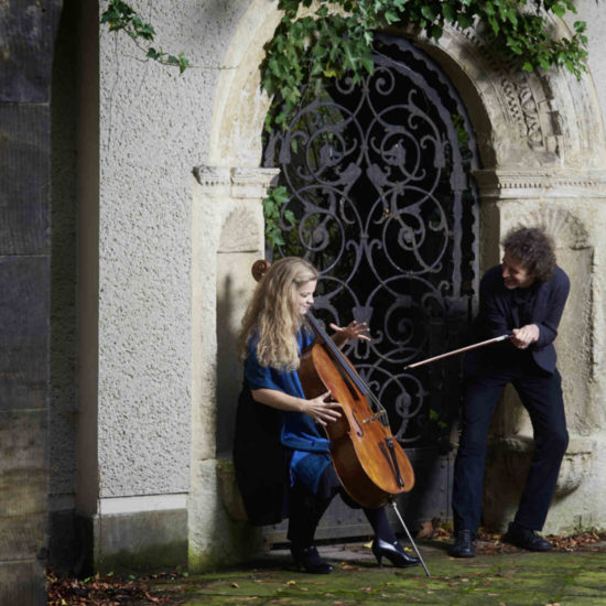 Die Cellistin Alma Stolte sitzt mit Ihrem Cello in einem Torbogen, daneben steht ihr Duo-Partner Pianist Friedemann Stolte und hält ihren Cellobogen