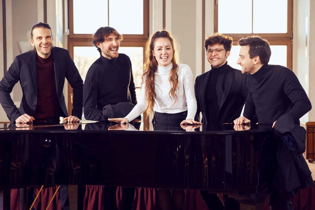 Die Leipziger Jazzband DÉJÀ VU steht an einem Flügel und lacht in die Kamera
