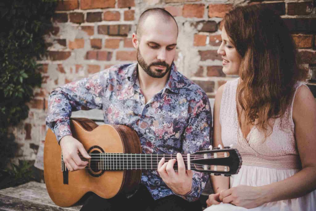 SARAH und LUCA sitzen vor einer Backsteinmauer und präsentieren Livemusik mit Gesang und Gitarre