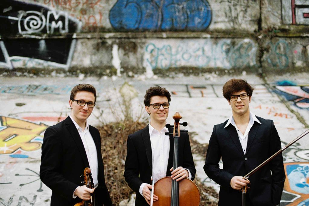 Zwei Geiger und ein Cellist lächeln in die Kamera