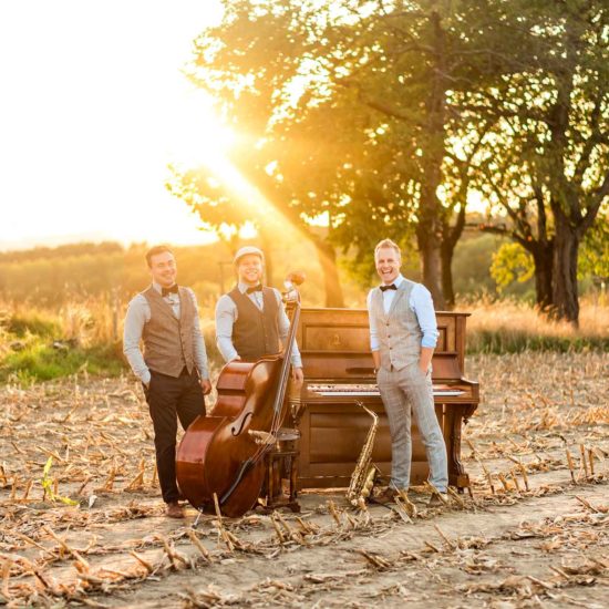 Ein Saxophonist, ein Pianist und ein Kontrabassist stehen auf einem Feld und lachen in die Kamera