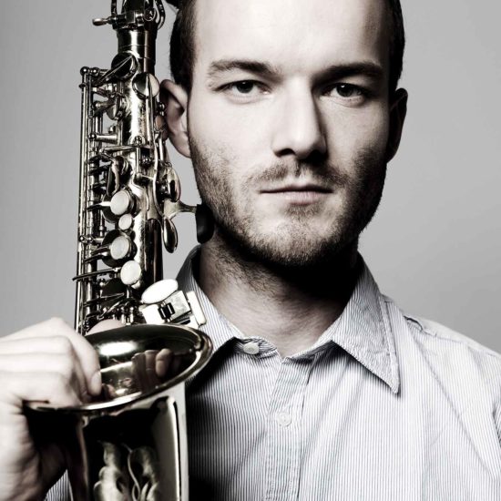 Der Saxophonist Richard Ebert hält sein Saxophon neben dem Kopf und schaut in die Kamera
