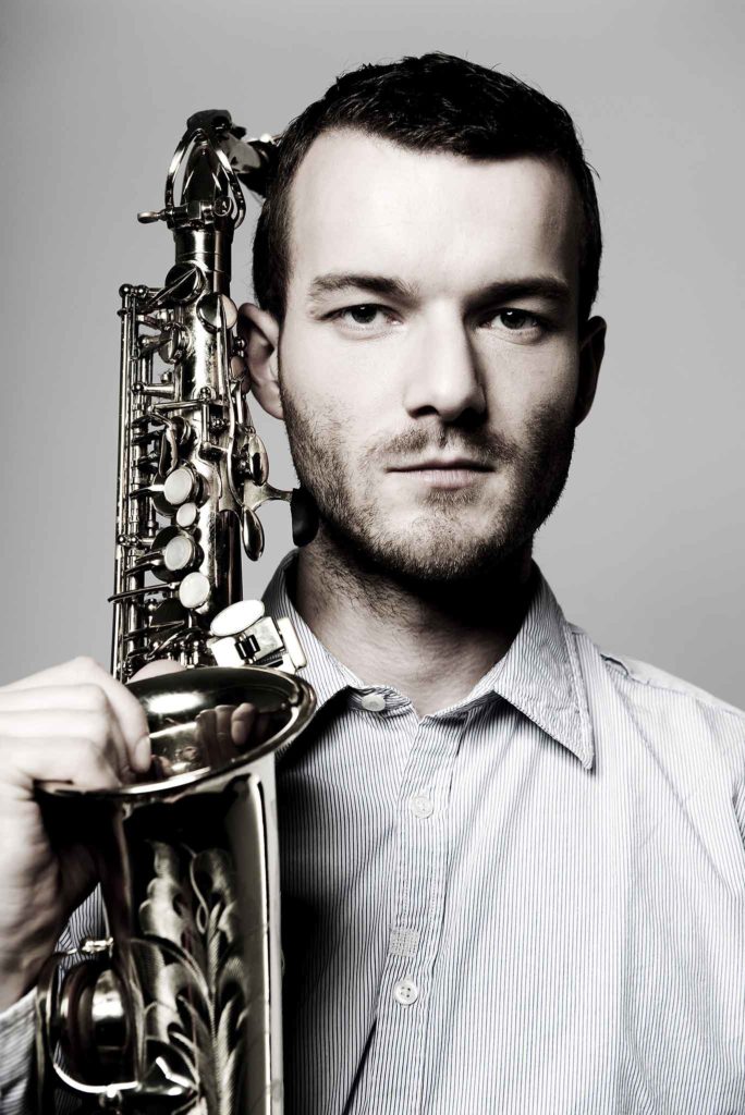 Der Saxophonist Richard Ebert hält sein Saxophon neben dem Kopf und schaut in die Kamera